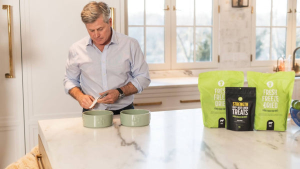 A dog owner prepares his dogs meals, with Get Joy's dog food product range displayed on a white marble countertop.