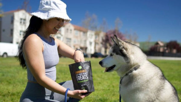 A woman wearing a bucket hat holds a black bag of Get Joy's superfood dog treats as her Husky dog patiently waits for one.
