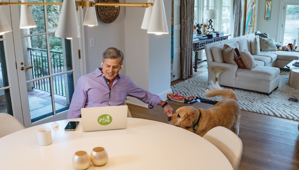 A woman holds up a black bag of Get Joy's Calm+ supplements and smiles at her dog, who is looking at the bag.