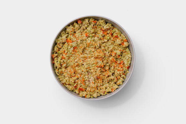 A white lab dog looks at a silver bowl of food that has dog food topper on it.