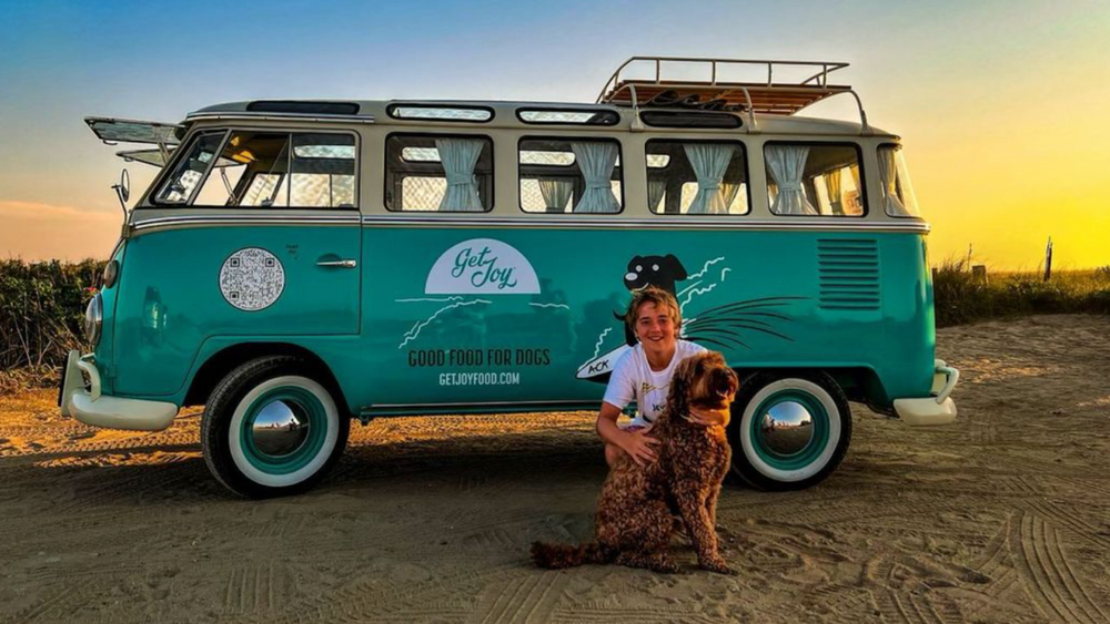 A dog and its owner pose in front of a teal vintage VW bus/