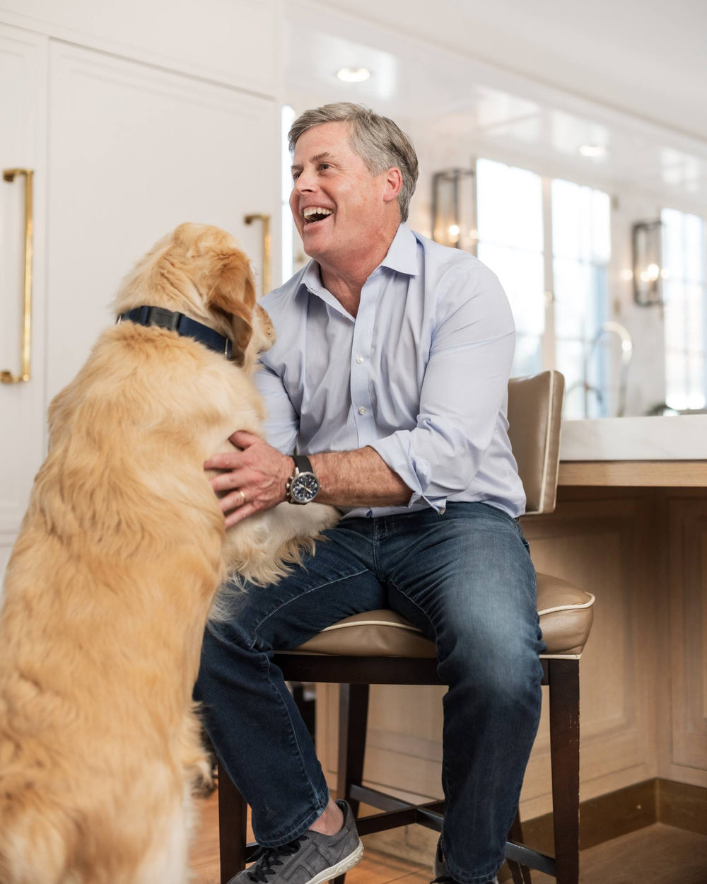 A man sitting on a kitchen stool while giving his dog a love tap.