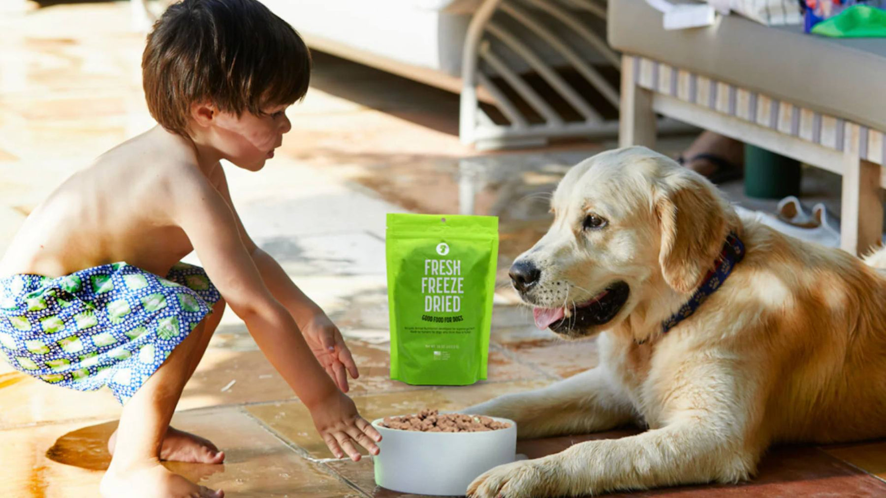 A young boy squats outside next to his dog, who is sitting next to a green bag of Get Joy fresh food.
