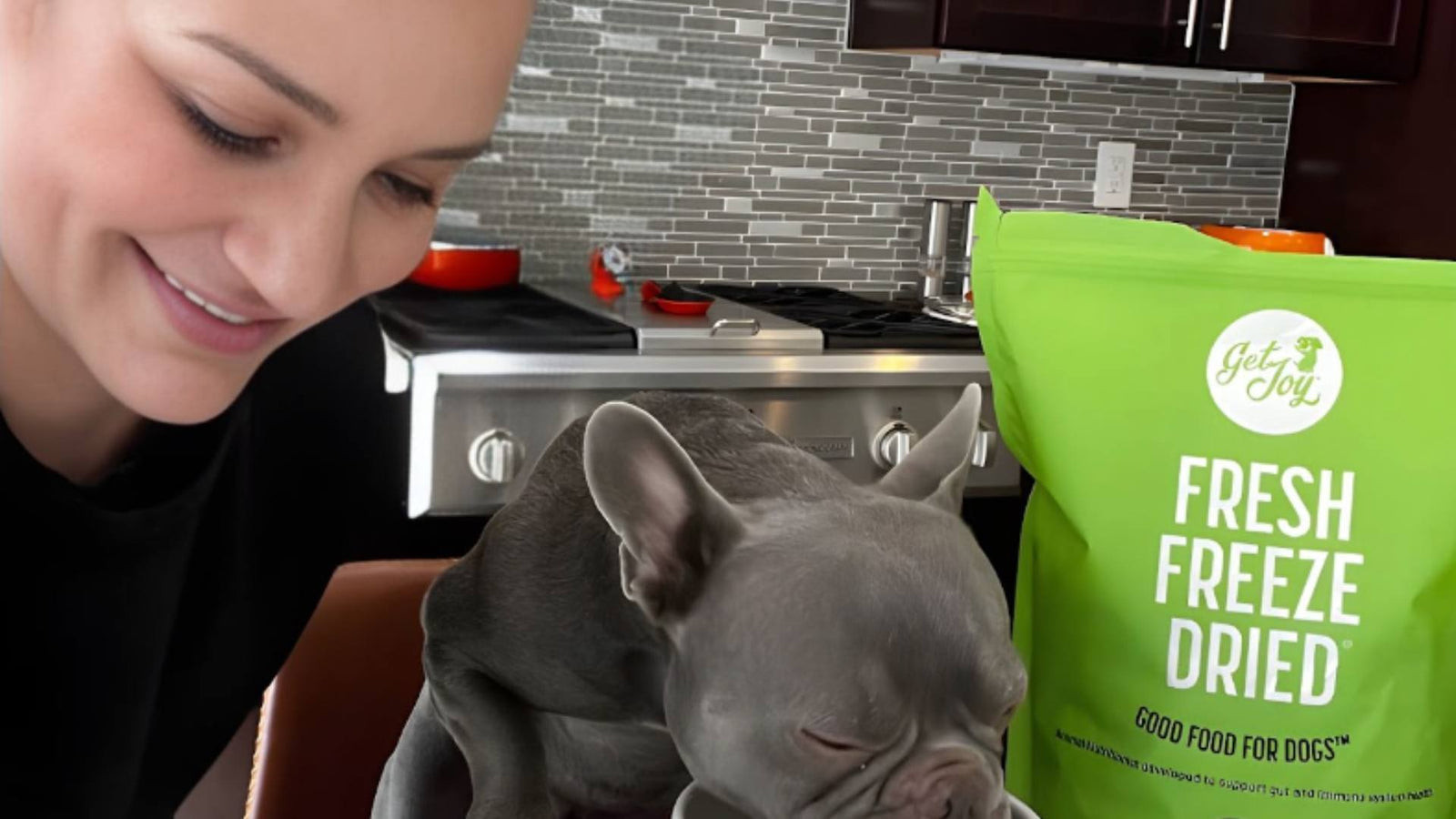 A woman smiles next to her dog, who sits and eats next to a green bag of Get Joy's fresh freeze dried dog food.