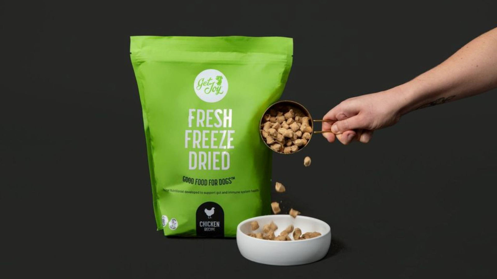 Against a black background, a green bag of Get Joy's fresh food for dogs is displayed. A person's arm is in the photo, pouring the fresh freeze dried food into a small white bowl.