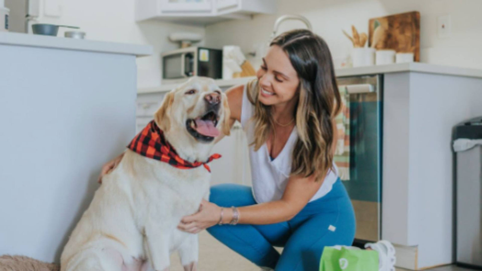 A woman kneels in the floor of her kitchen next to a bag of green Get Joy food while petting her dog, demonstrating dog gut health.