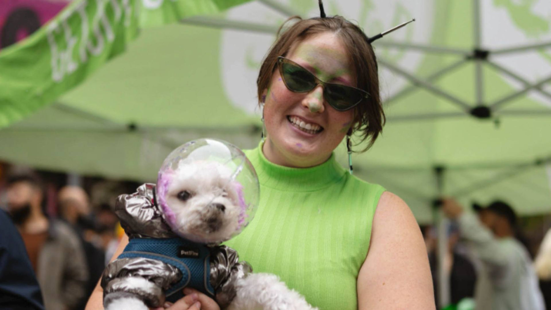 A woman wears a green tank top and sunglasses while holding her fluffy white dog, who's wearing a space costume.