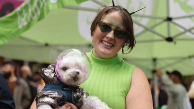 A woman wears a green tank top and sunglasses while holding her fluffy white dog, who's wearing a space costume.