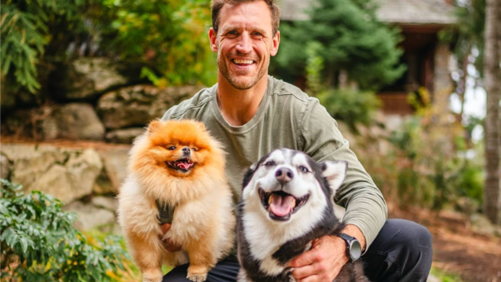 A person sits with two dogs outside, happily smiling. 