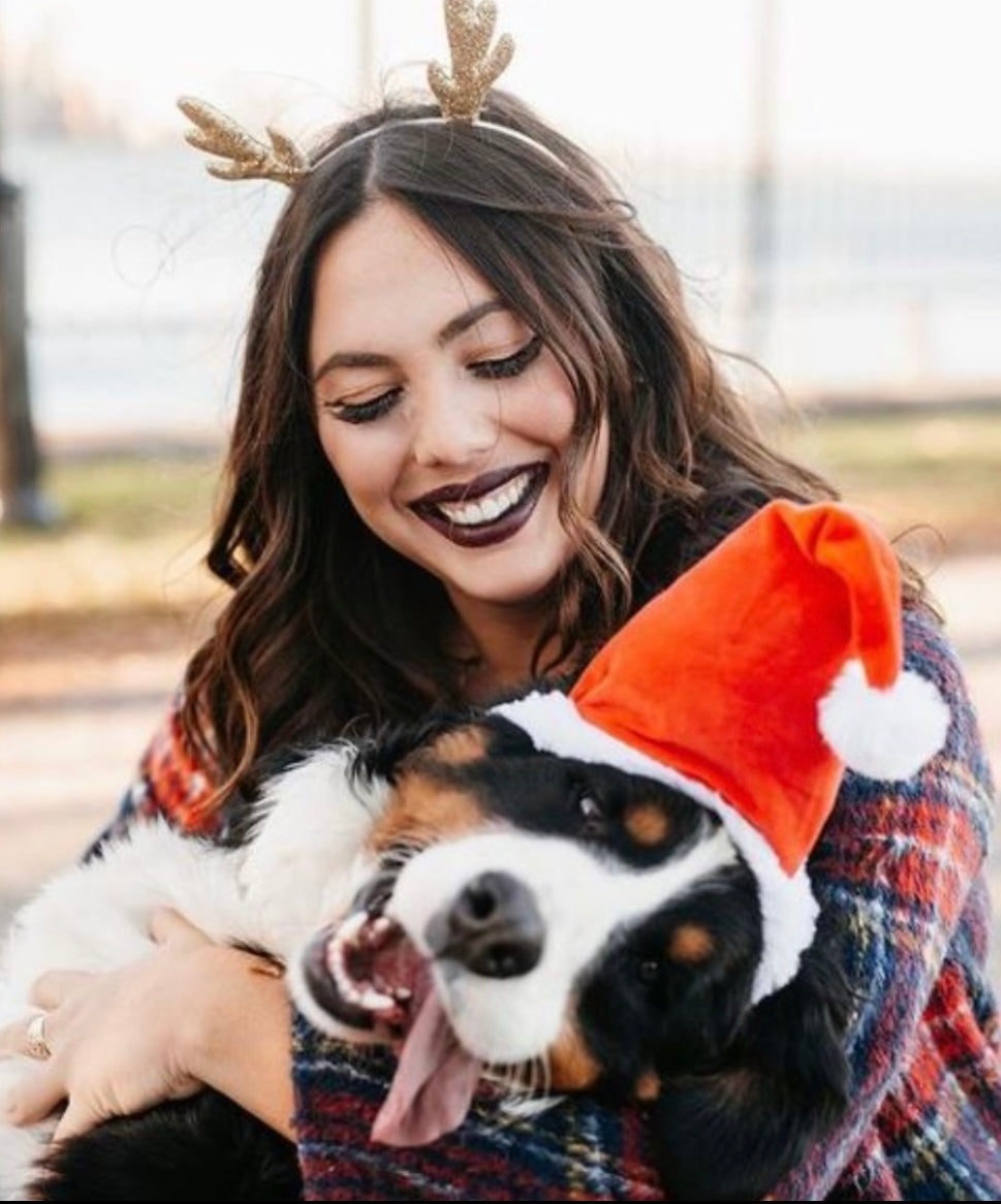 A young woman in reindeer antlers smiles down at her goofy dog in a Santa hat that he got in a dog advent calendar.