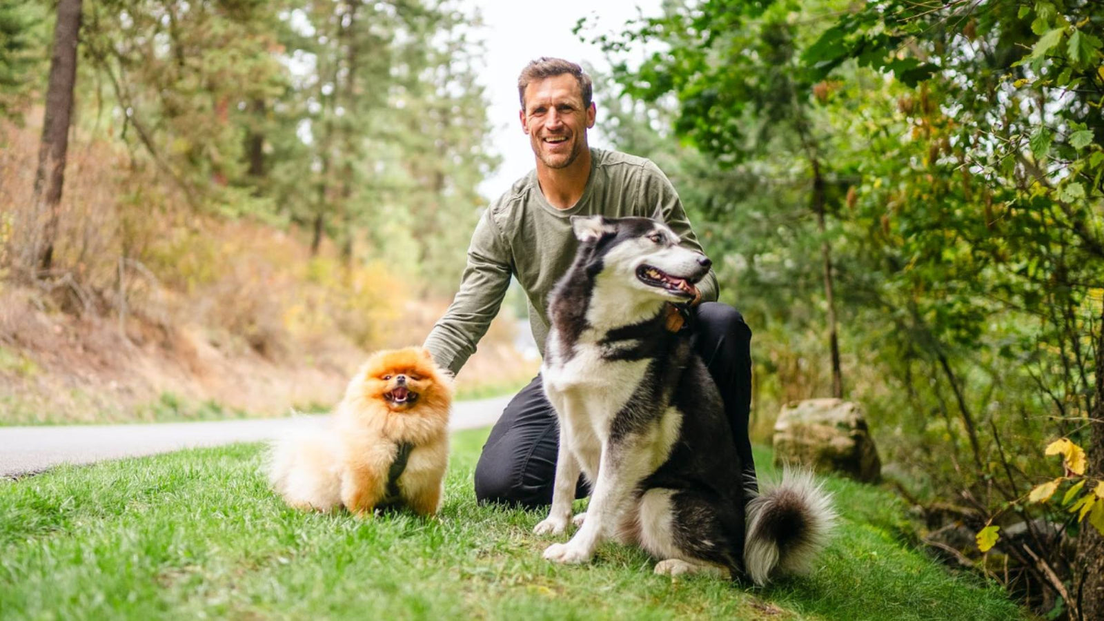 A man smiles and poses outside with a small fluffy dog and a Husky dog.