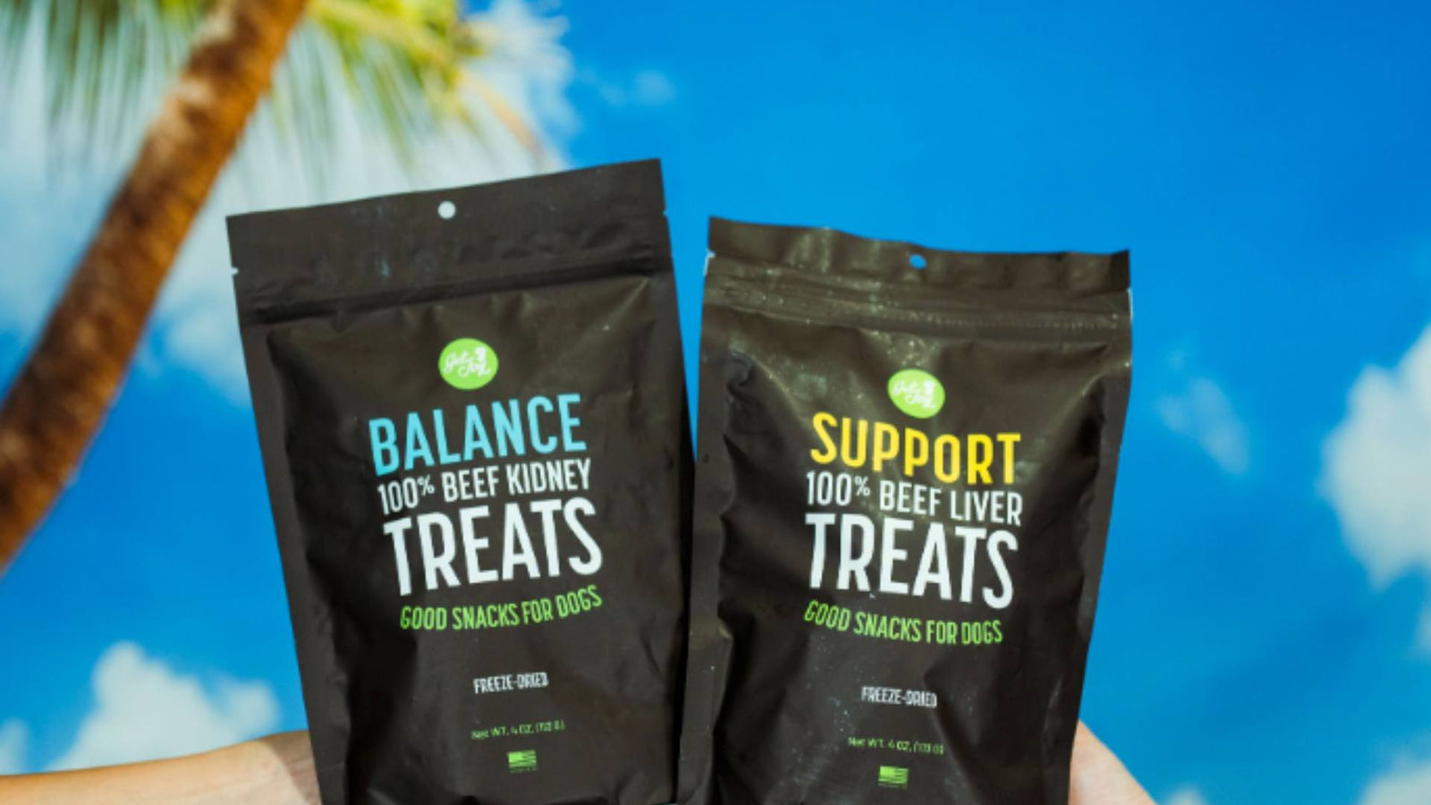 Two black bags of Get Joy's dog treats are displayed in front of a blue sky and palm tree background, demonstrating enhancing nutrition for dogs.