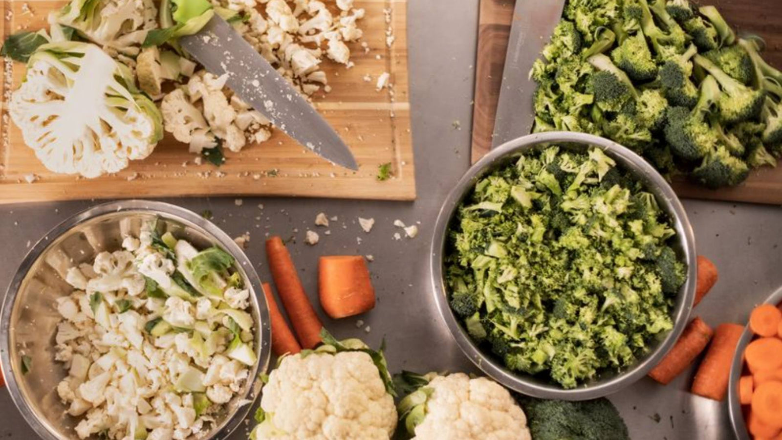 Vegetables like broccoli, cauliflower, and carrots are displayed in bowls and on a wooden cutting board.