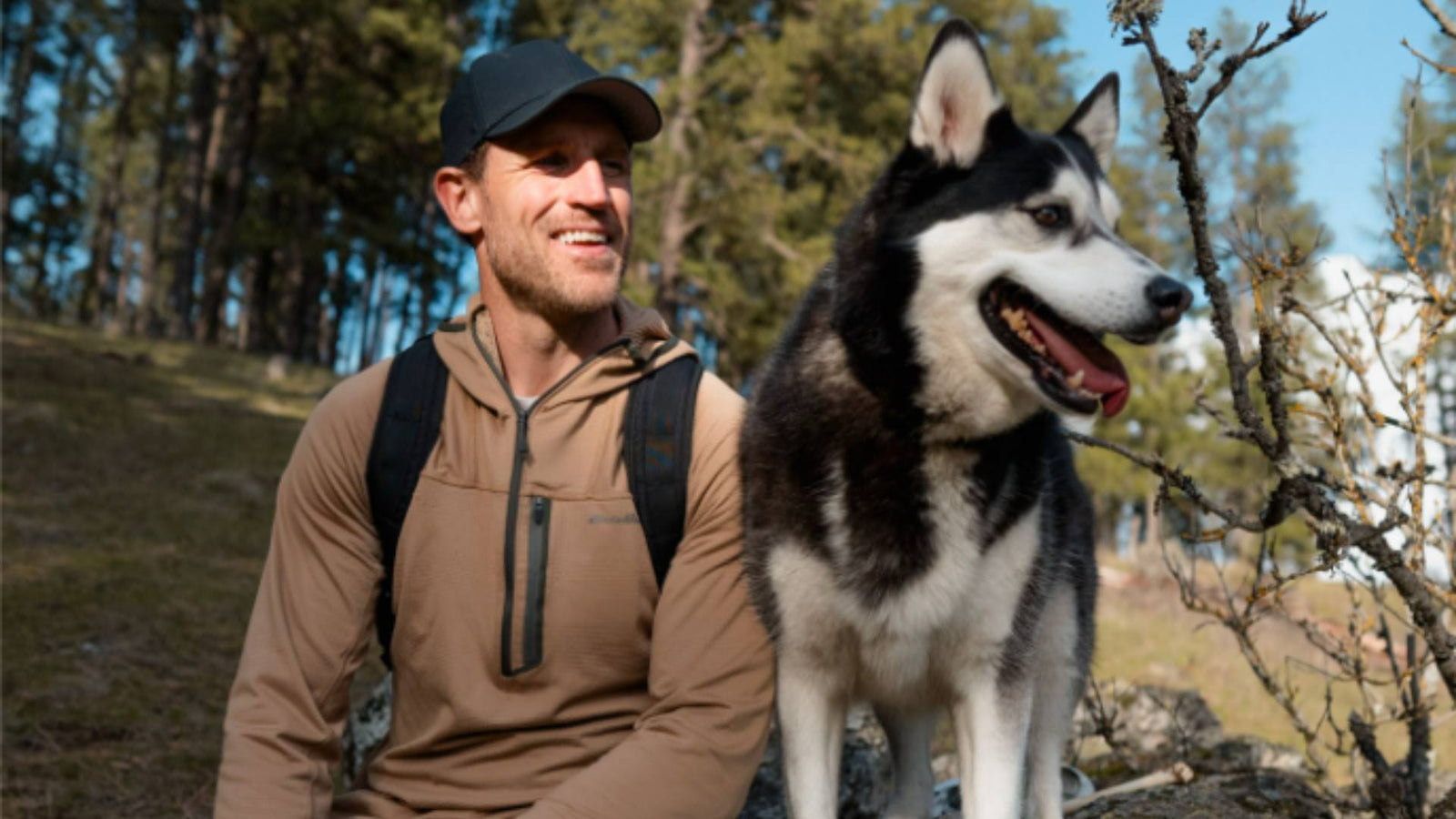 A man sits on a rock with his Husky dog and smiles as they take in the sunny nature around them, demonstrating a successful hiking trip with his dog.