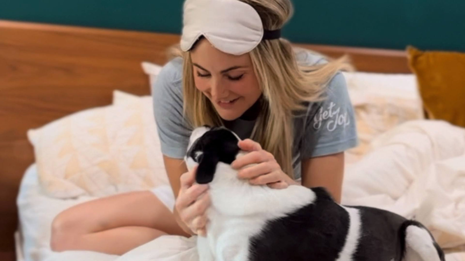 A woman wears pajamas and an eye mask on her head while sitting in bed and smiling, holding her dog. She is demonstrating providing a calm environment for dog anxiety.