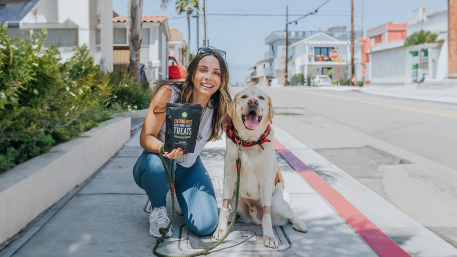 A woman holds a black bag of Get Joy's dog treats while kneeling next to her dog, a lab.