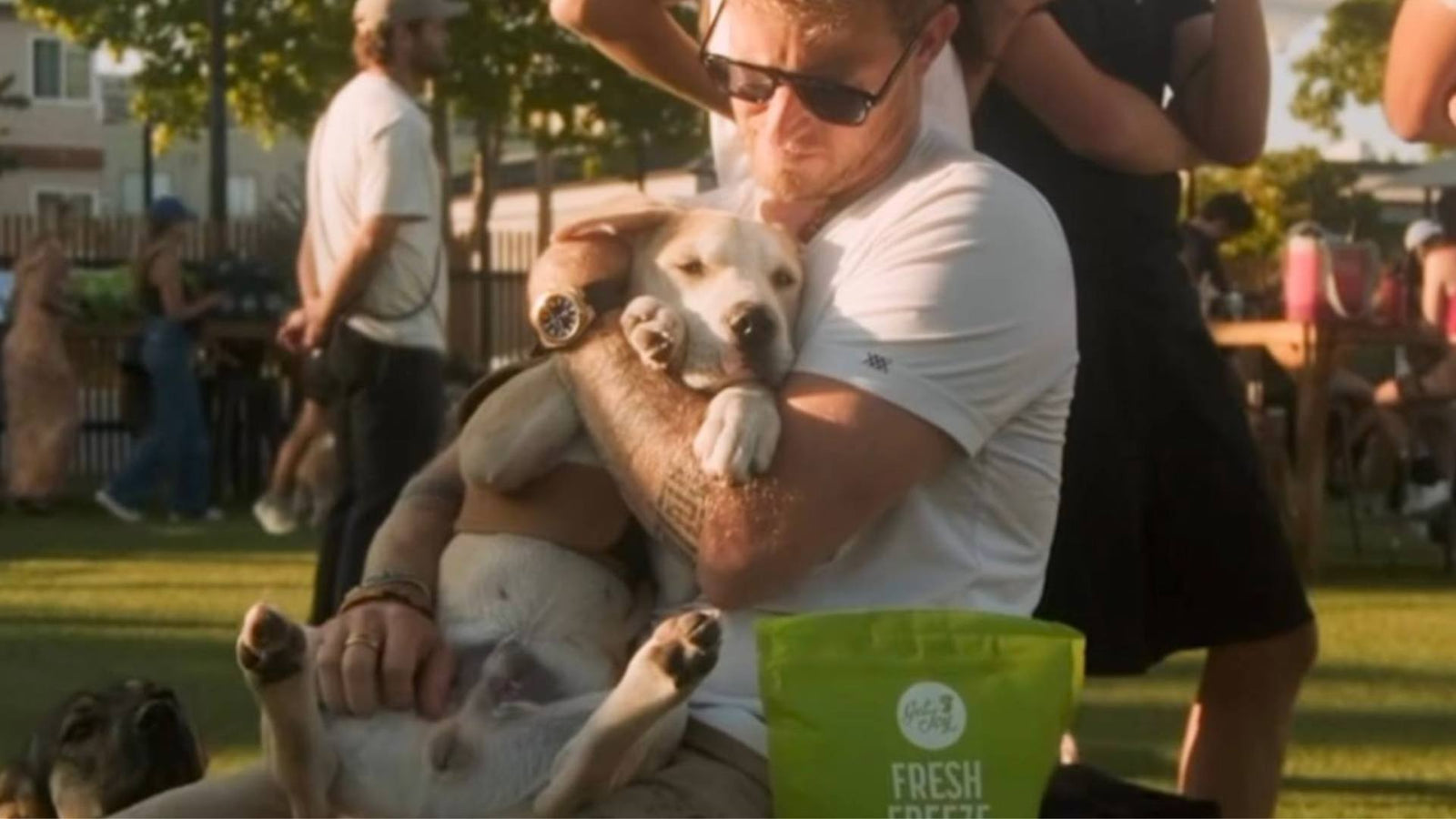 A man sits outside wearing black sunglasses and holds a dog in his arms next to a green bag of Get Joy dog treats, demonstrating successful pet and dog adoption.