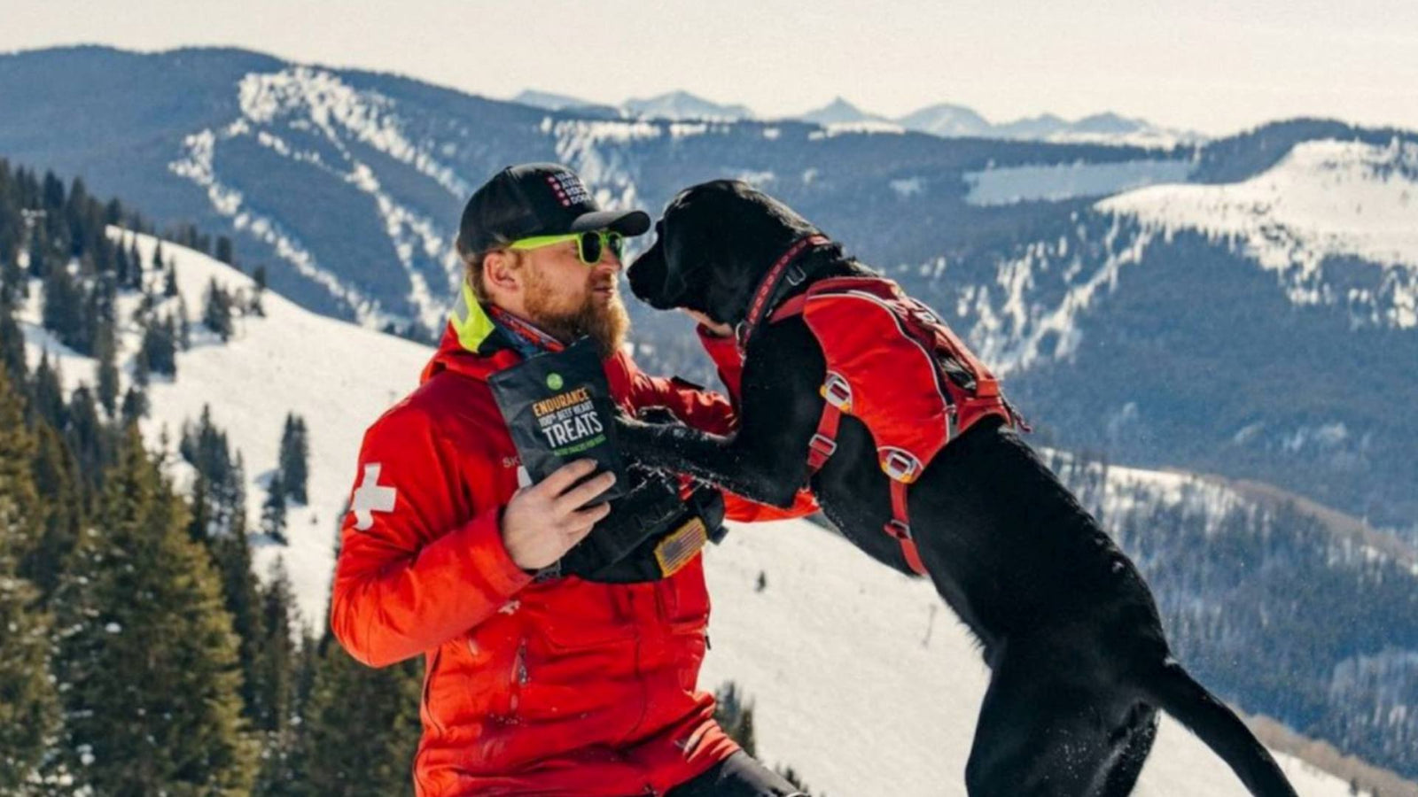 A person wearing a red emergency jacket stands outside on a mountain in the snow holding a bag of Get Joy's dog treats. A Black dog is wearing a red vest and standing on the human.