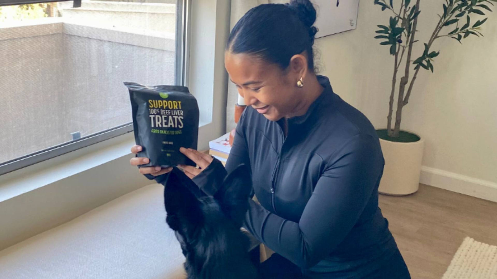 A woman sits next to her window in front of a green plant in a white pot and holds up a black bag of Get Joy's dog treats to her black dog's face.