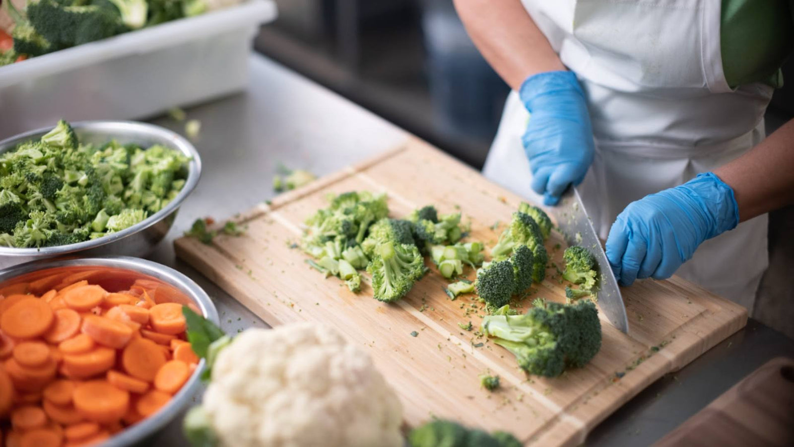 A person stands at a wooden cutting board while wearing blue gloves, cutting vegetables like broccoli and carrots, demonstrating pet wellness through nutrition.