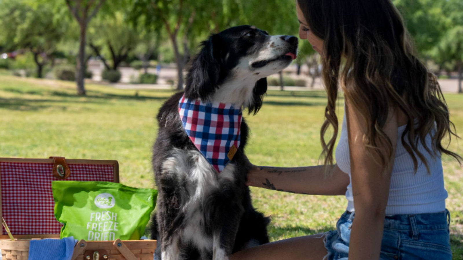 A dog wearing a checkered bandana around his neck sniffs his owners nose and he smiles and looks at him. A green bag of Get Joy's fresh freeze dried food is displayed, demonstrating eco friendly dog products.