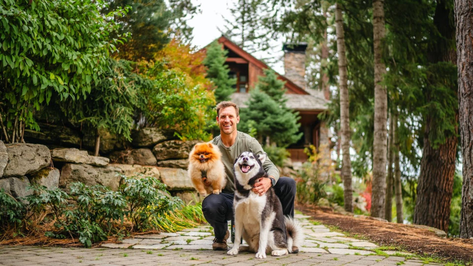 A man kneels outside while holding one small, tan colored puppy. A Husky dog sits next to him on his other side. The background displays a cabin in a lush forest.