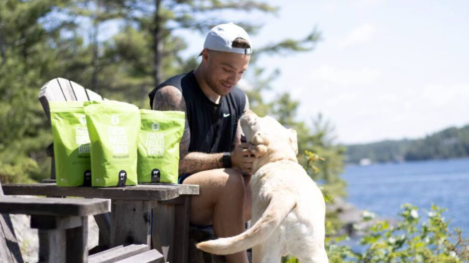 A dog dad wears a black tank top and a gray backwards hat while sitting in a picnic chair next to a blue lake. He is holding his dogs face and three green bags of Get Joy's dog food is displayed.