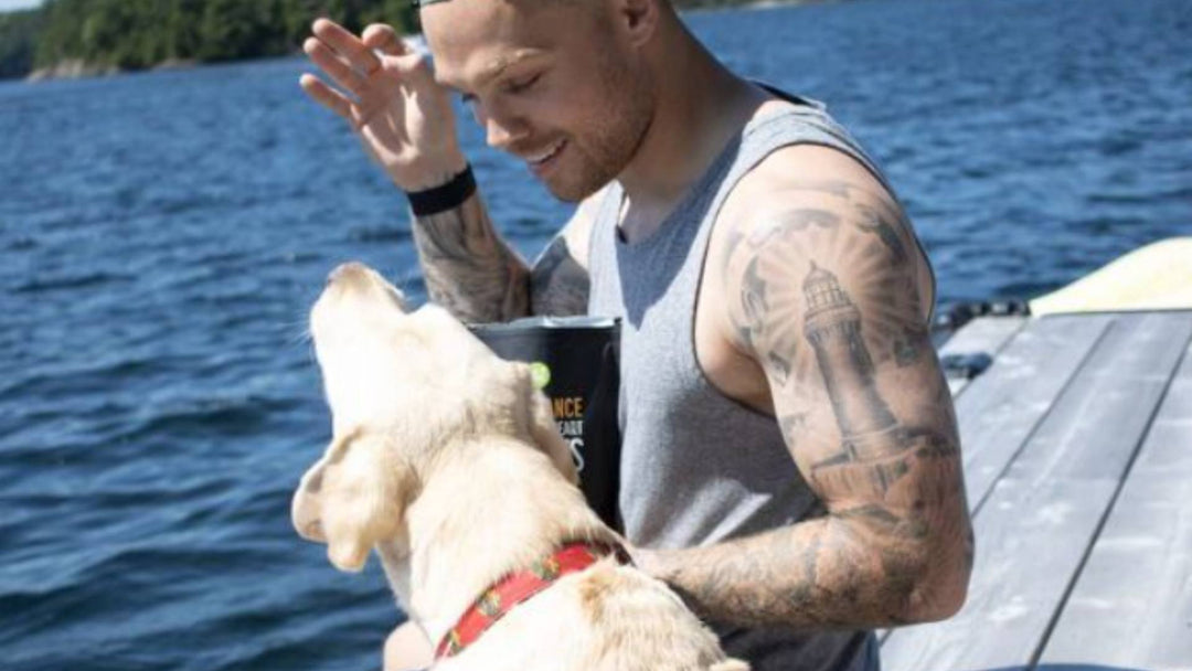 A man smiles and holds a dog treat up as his dog sits next to him and looks up at it.